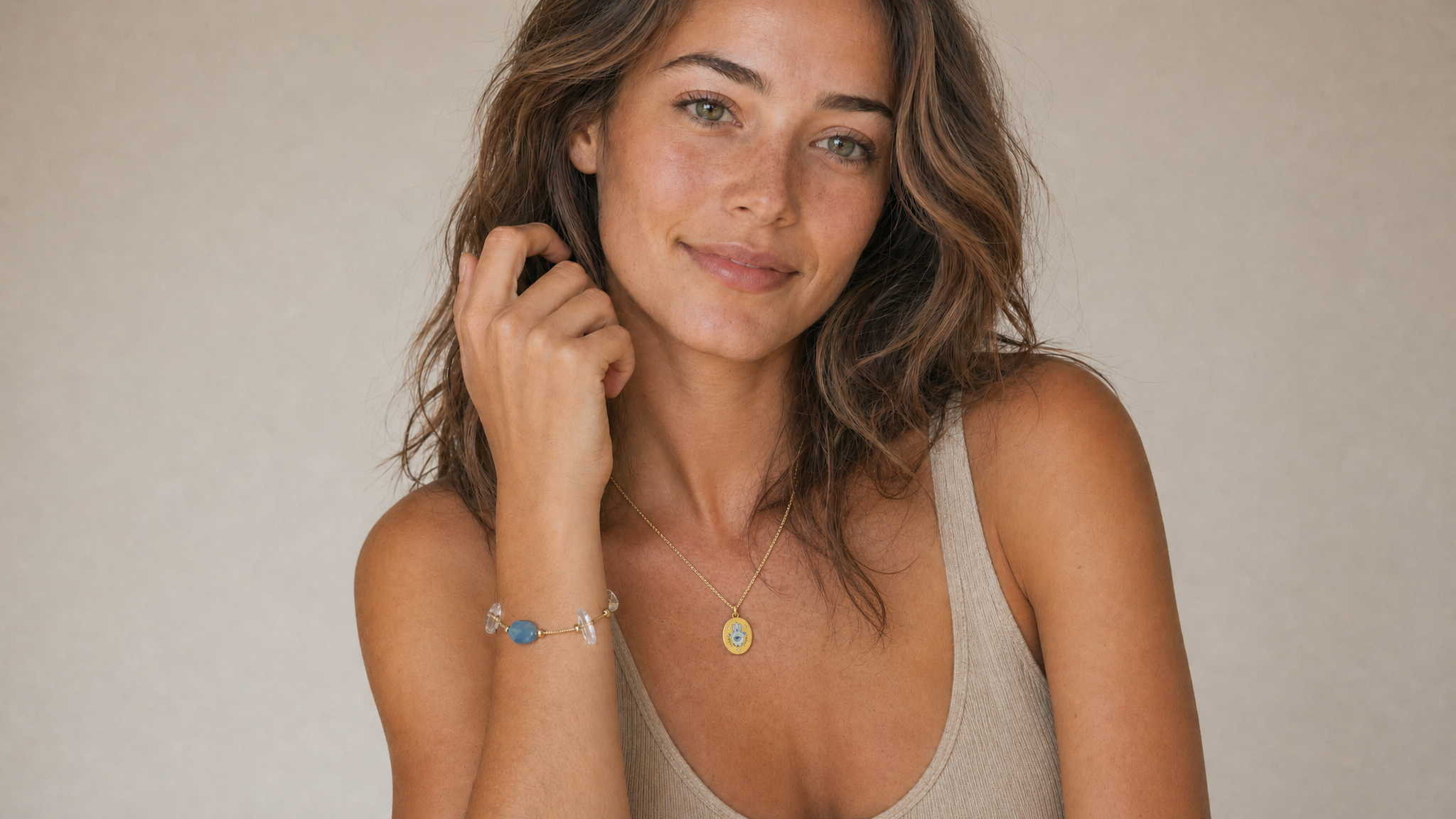 A young woman in a beige tank top smiling at the camera, wearing a Peace Piece Seren aquamarine bracelet on her wrist and an Elysian hamsa hand-painted enamel pendant at her collarbone