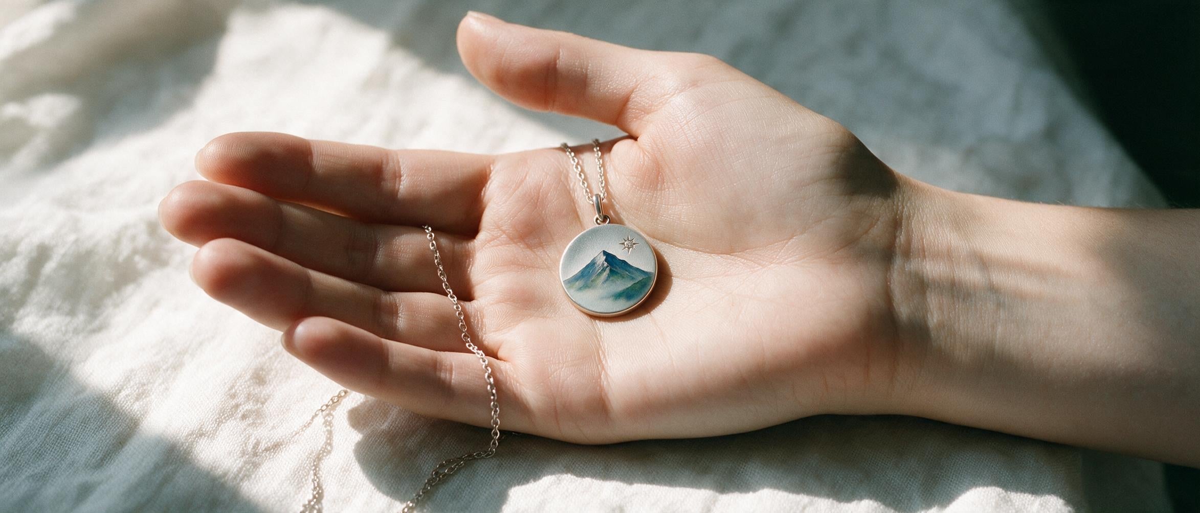 A silver enamel pendant painted with a mountain peak, held in an open palm